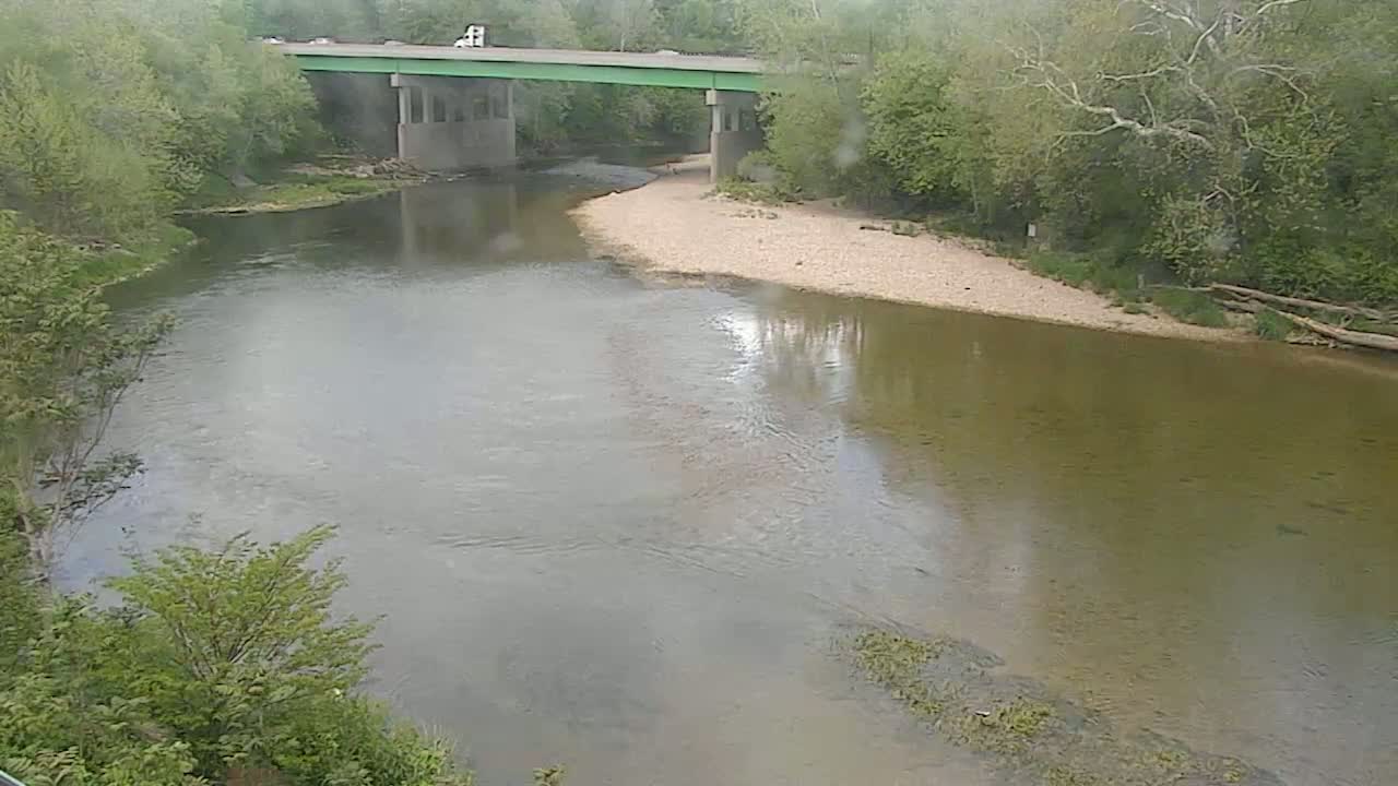 A murky river flows under a green bridge, bordered by lush green trees in rainy weather.