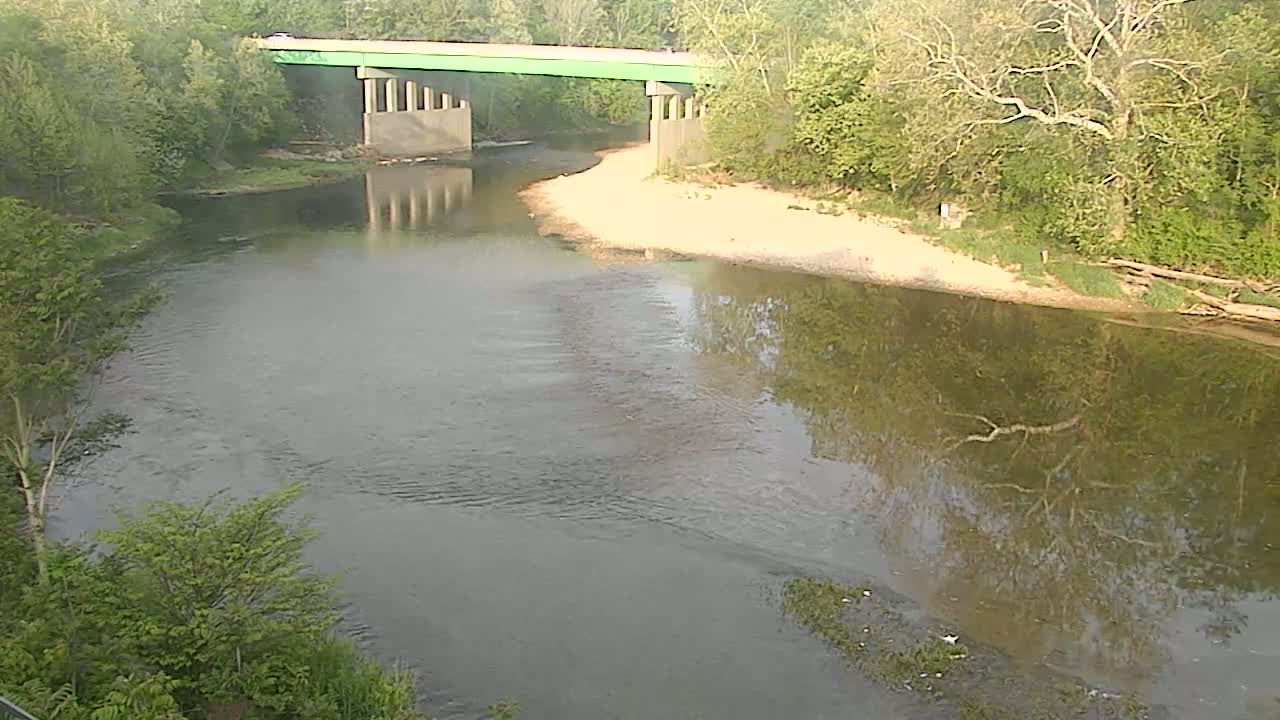 A river flows under a bridge, surrounded by lush green trees on a partly sunny day.