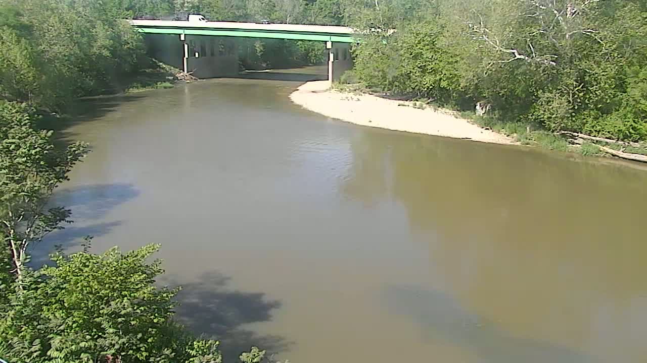 A murky river bends beneath a bridge surrounded by lush green trees on a sunny day.