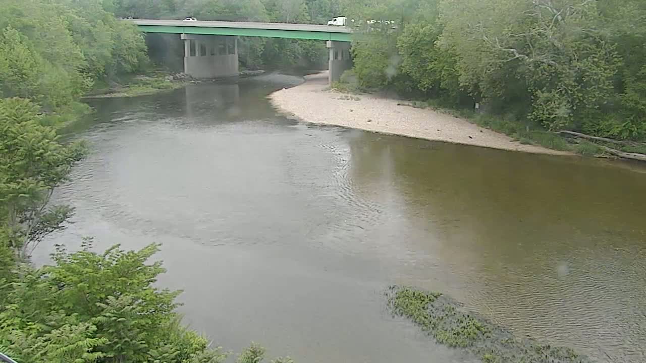 A river bends gently under a bridge on a rainy day, surrounded by lush green trees.