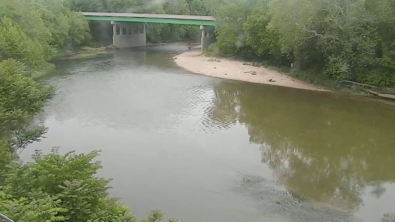 A calm, murky river flows under a bridge on a cloudy day, surrounded by lush green trees.