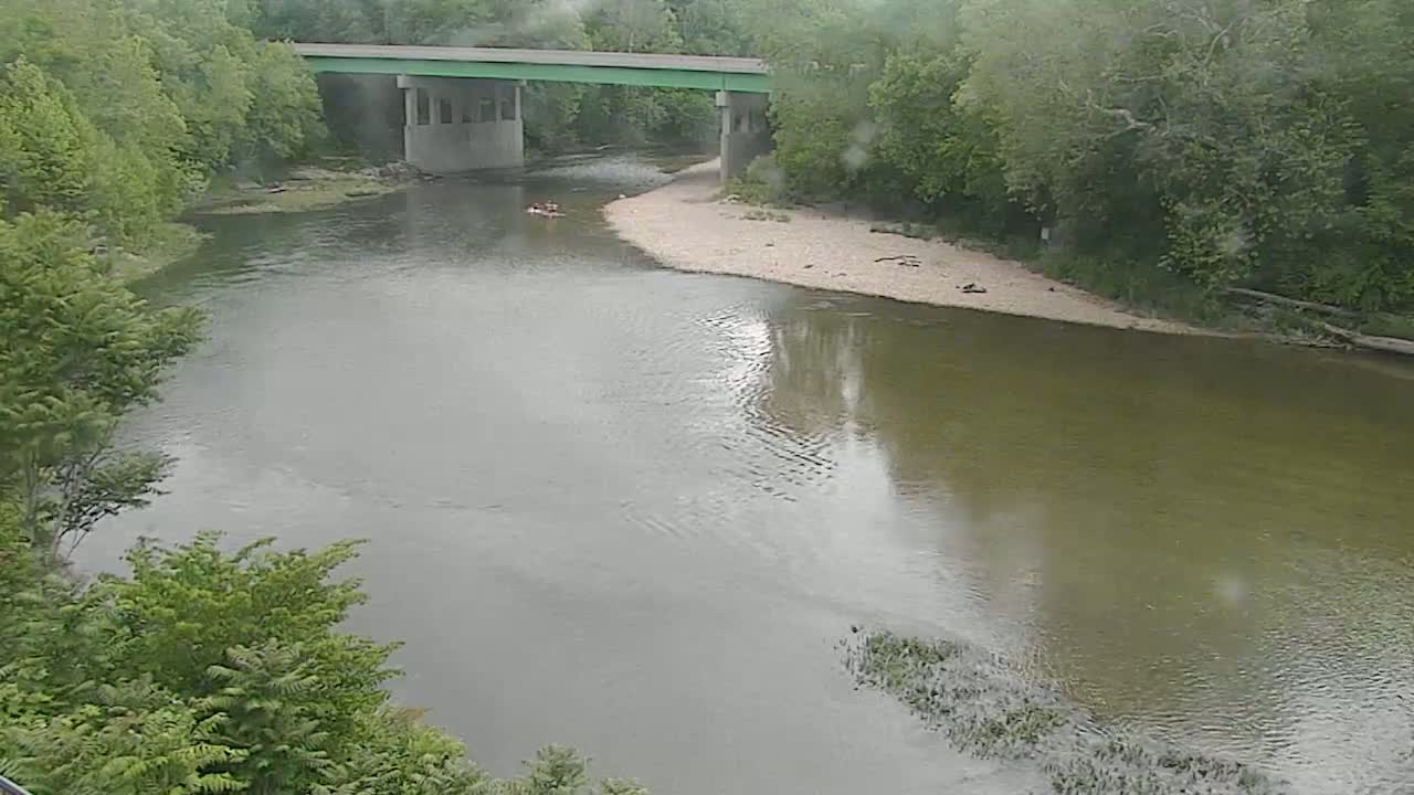 A river flows under a bridge on a rainy day, with several people visible in small boats on the water.