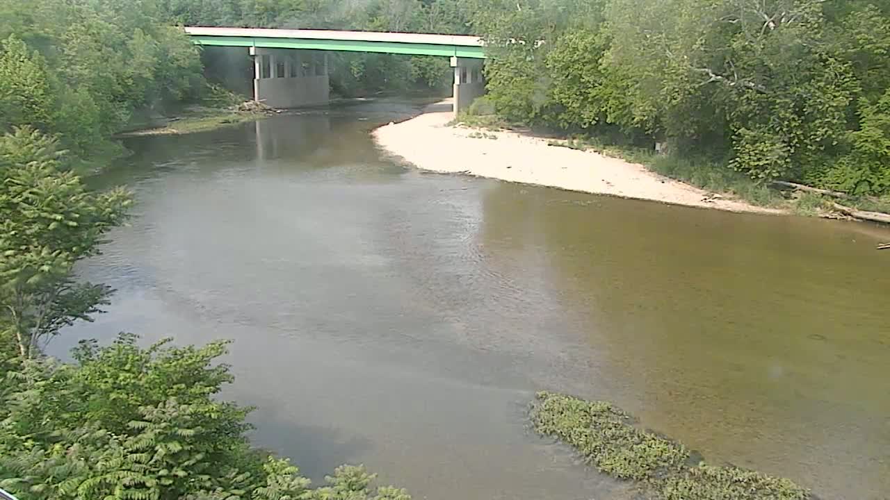 A light-green bridge spans a calm, light-brown river bordered by lush green trees under a seemingly clear sky.