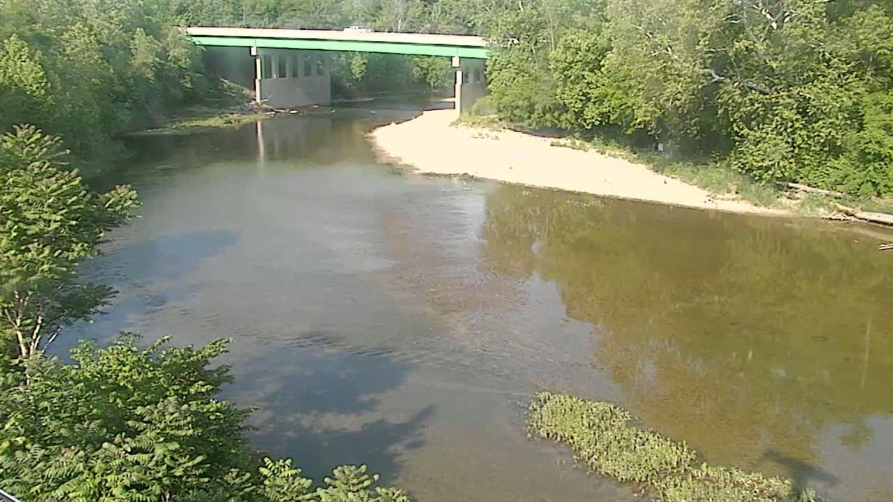 A calm river flows under a bridge surrounded by lush green trees on a sunny day.
