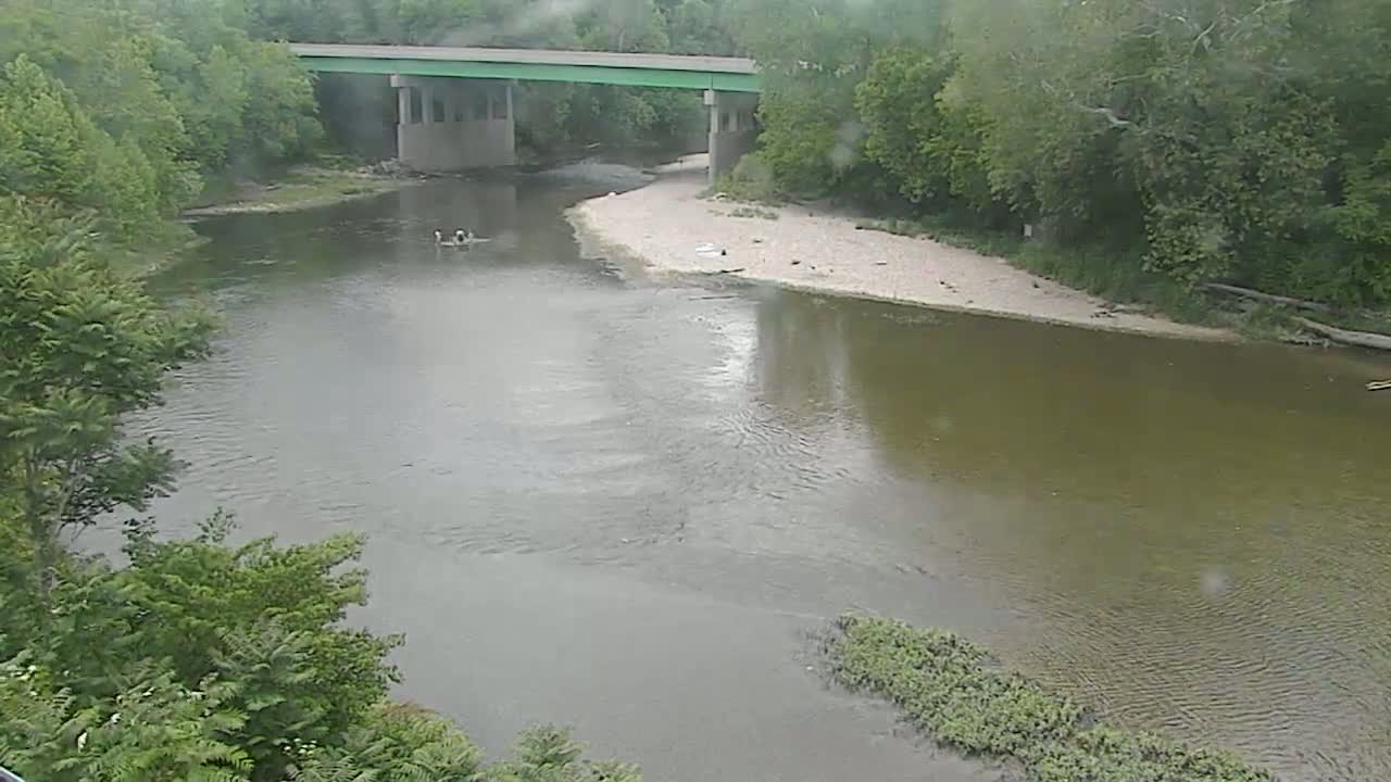 A river flows under a bridge on an overcast day, with people visible in the water near the riverbank.