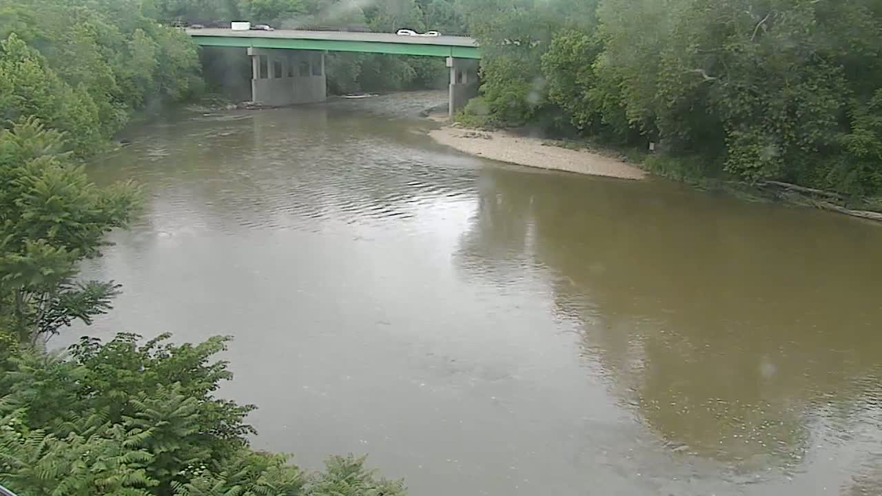 A muddy river flows under a bridge during a light rain, surrounded by lush green trees.
