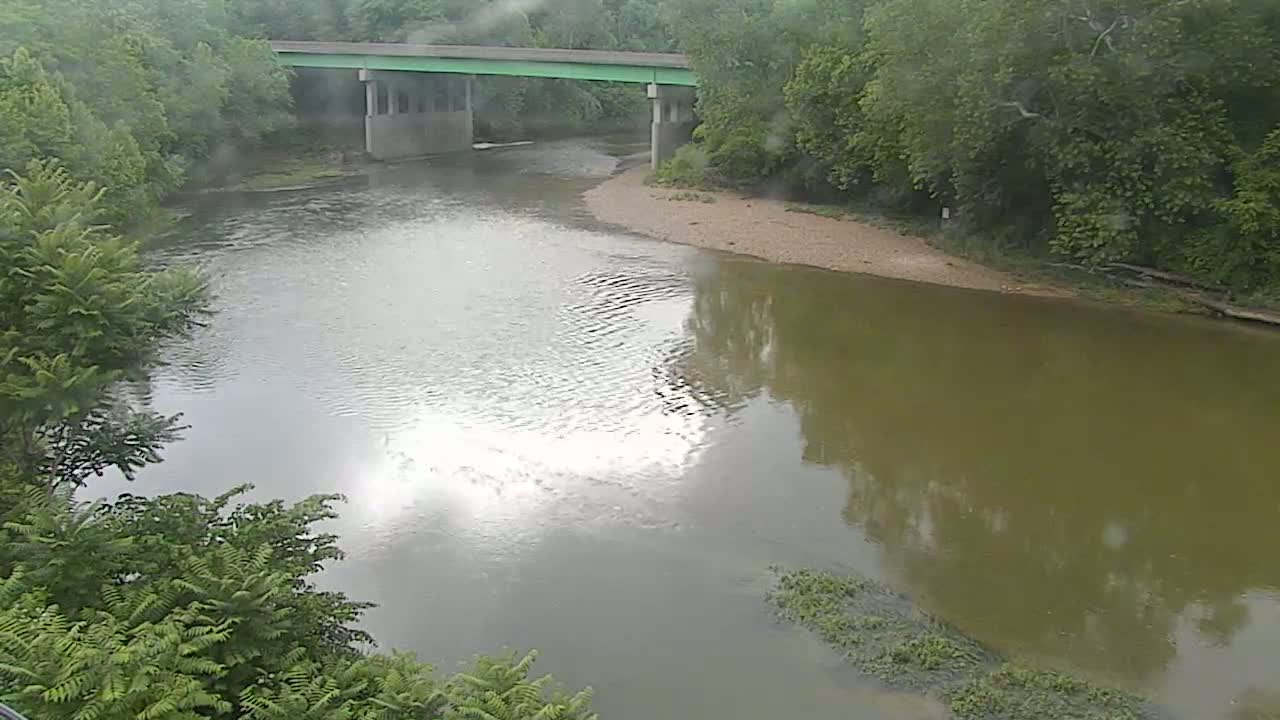 A river flows under a bridge on a cloudy day, surrounded by lush green trees.