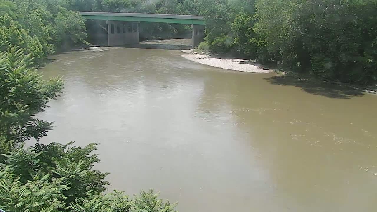 A wide, muddy river flows under a bridge surrounded by lush green trees on a cloudy day.