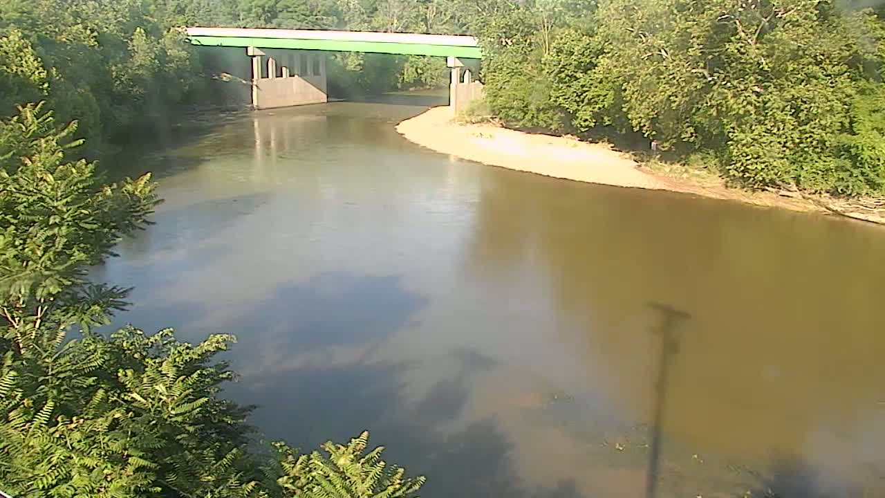 A light-green bridge spans a murky river bordered by lush green trees under a mostly clear sky.