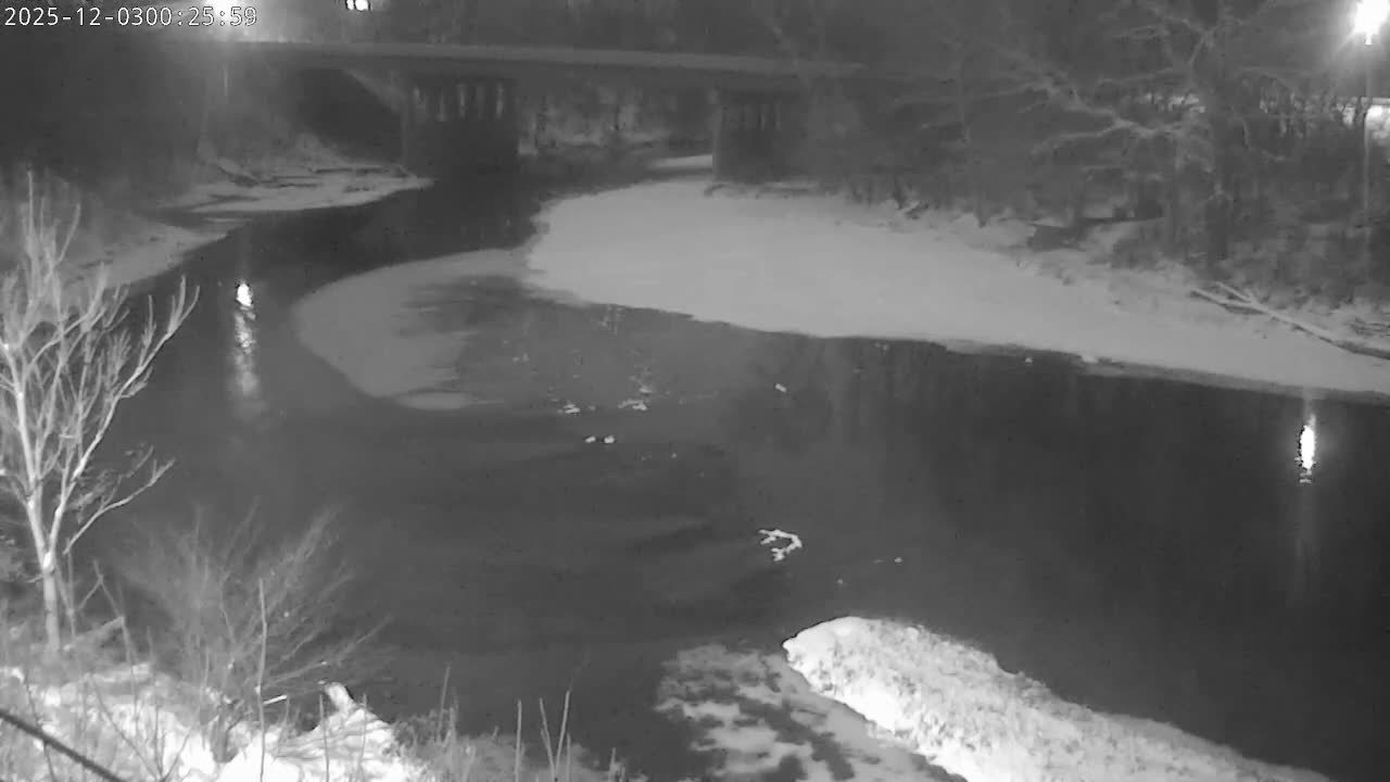 At night, a snow-covered landscape features a river partially frozen with ice, bordered by bare trees and crossed by a bridge in the distance, indicating cold, snowy conditions.