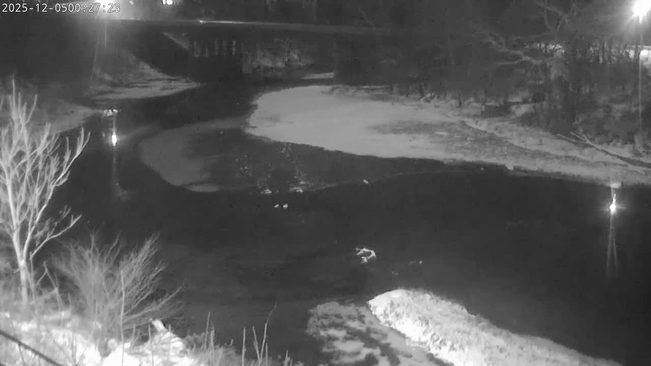 A monochrome nighttime image captures a winding river partially frozen with snow-covered banks, illuminated by scattered artificial lights under clear, cold conditions.