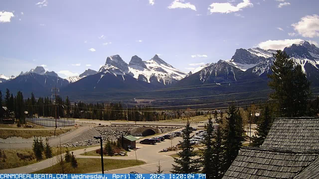 A partly sunny day reveals a snow-capped mountain range in the background, overlooking a parking lot, a small building, and some green spaces in the foreground.