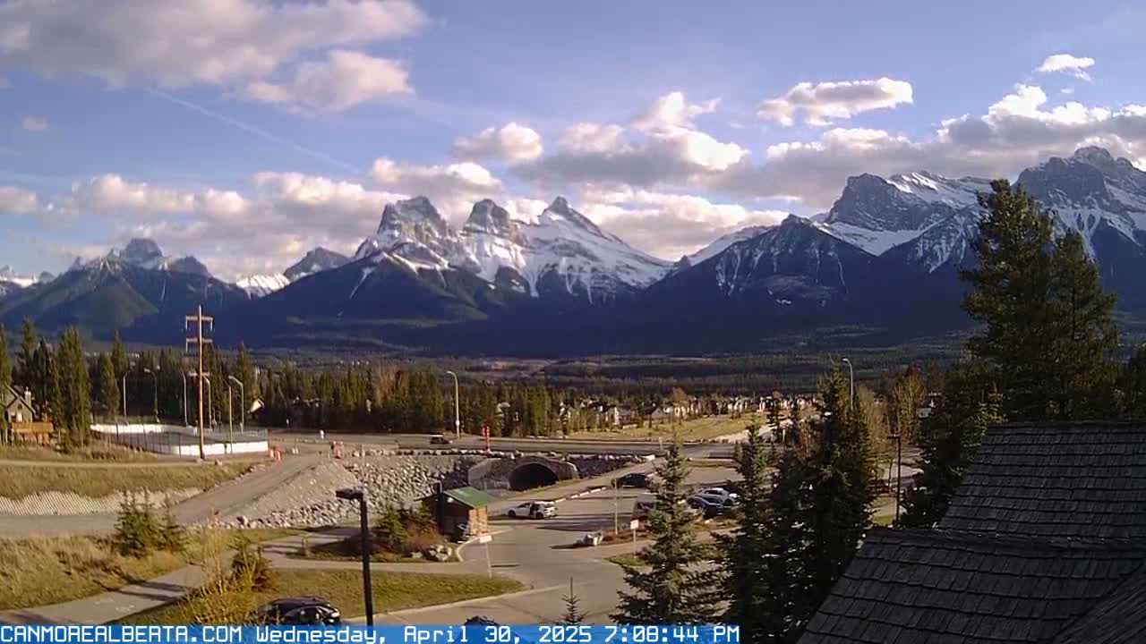 A partly cloudy afternoon view of snow-capped mountains in the distance, overlooking a valley with roads, buildings, and a parking area.
