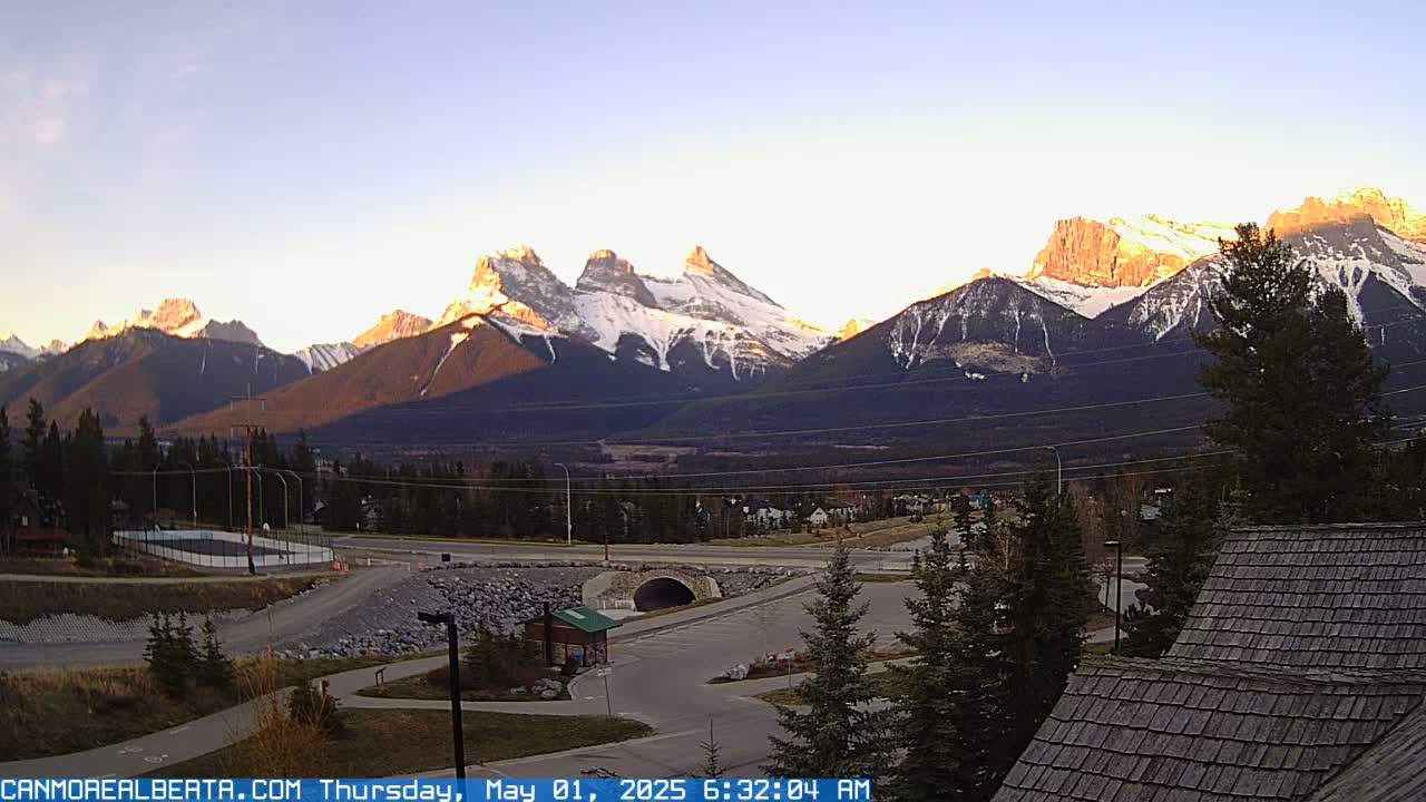 A sunrise illuminates snow-capped mountains behind a town with a road, parking lot, and a small tunnel.