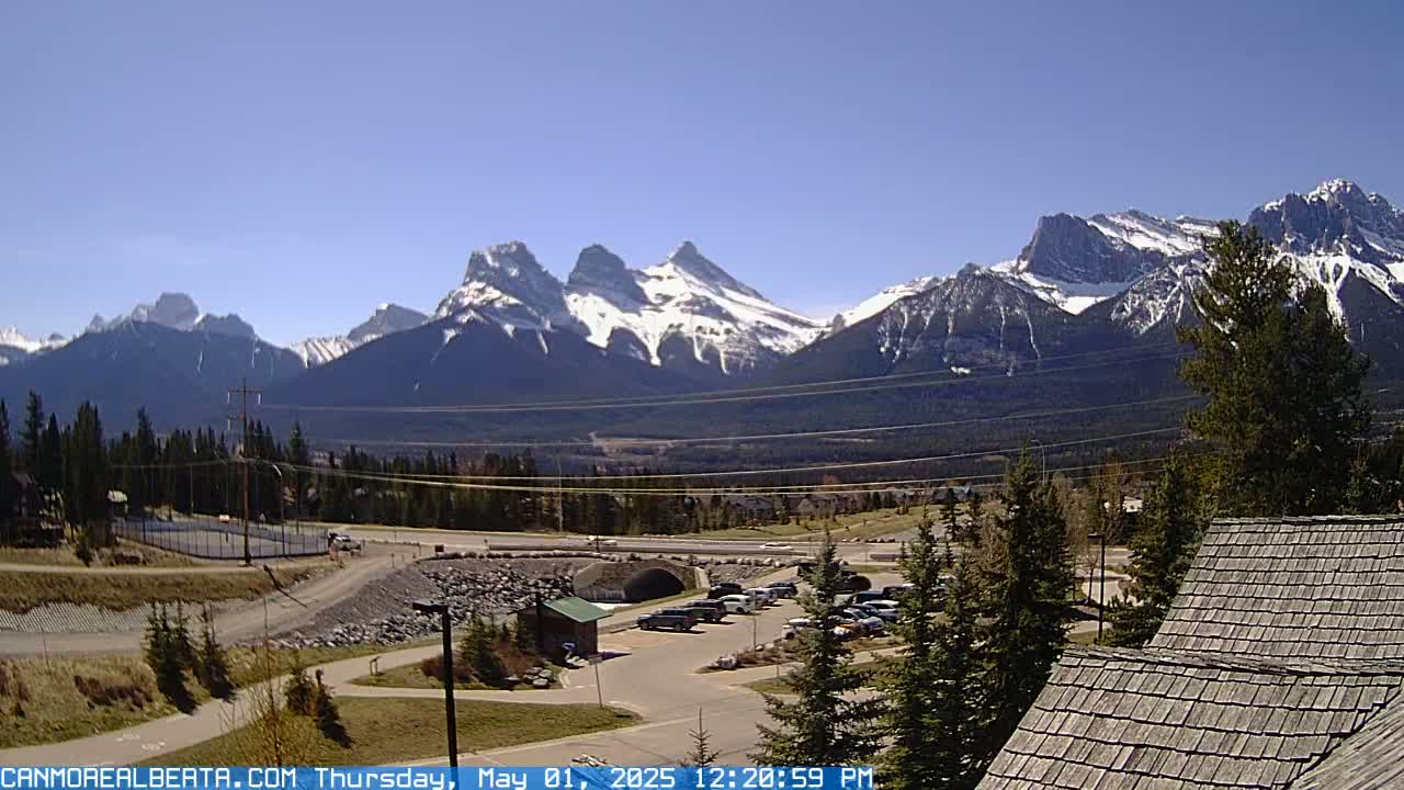 A snow-capped mountain range is visible in the background on a clear, sunny day, overlooking a parking lot, road, and some buildings.