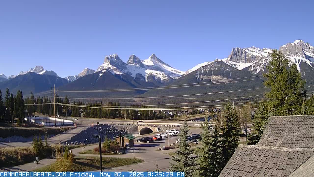 A sunny day reveals snow-capped mountains in the background, overlooking a valley with a road, parking lot, and some buildings in the foreground.