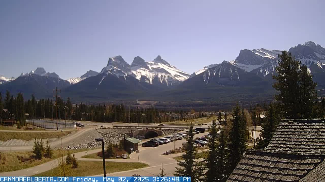 A snow-capped mountain range is visible in the distance on a clear, sunny day, overlooking a parking lot, some trees, and a road.