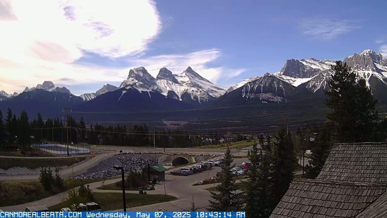 A partly cloudy day reveals snow-capped mountains in the distance, overlooking a town with a parking lot and a small bridge.