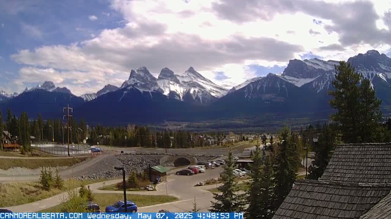 A partly cloudy day reveals snow-capped mountains in the distance, overlooking a valley with a parking lot, some buildings, and green spaces.
