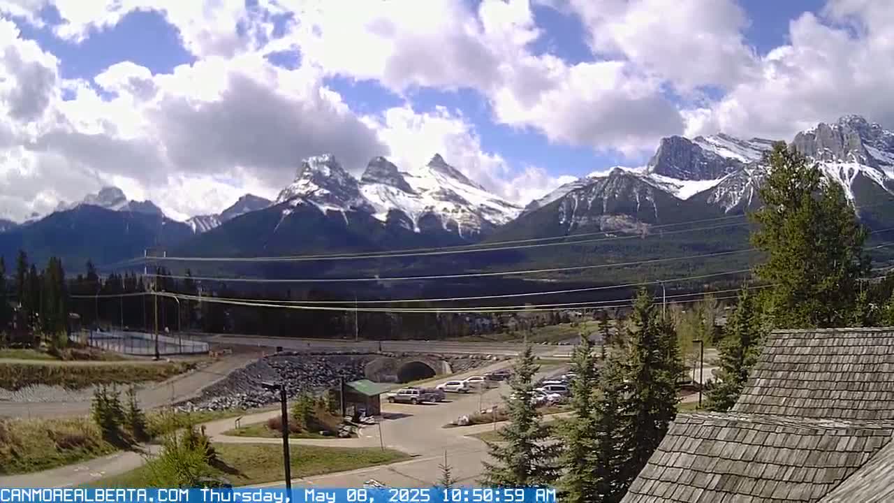 A partly cloudy day reveals snow-capped mountains in the distance, overlooking a parking lot, roadway, and a small building with a wood shake roof.