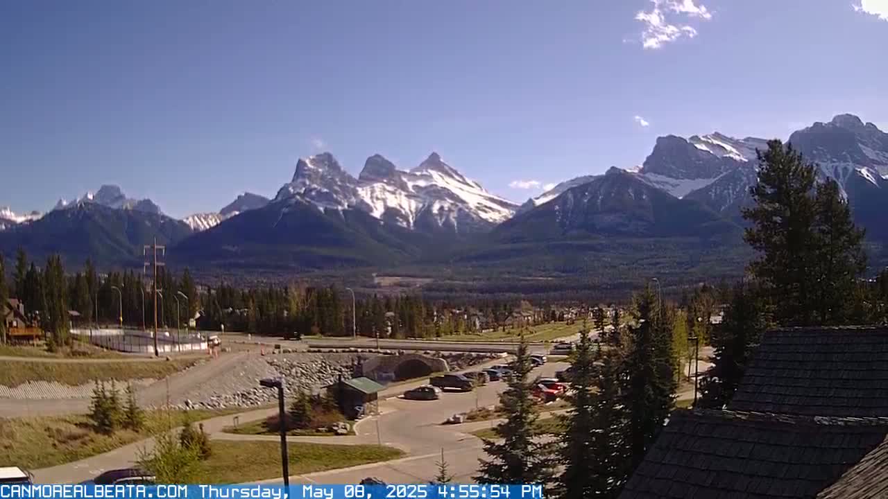 A partly cloudy day reveals snow-capped mountains in the distance, overlooking a valley with a parking lot, some buildings, and evergreen trees in the foreground.