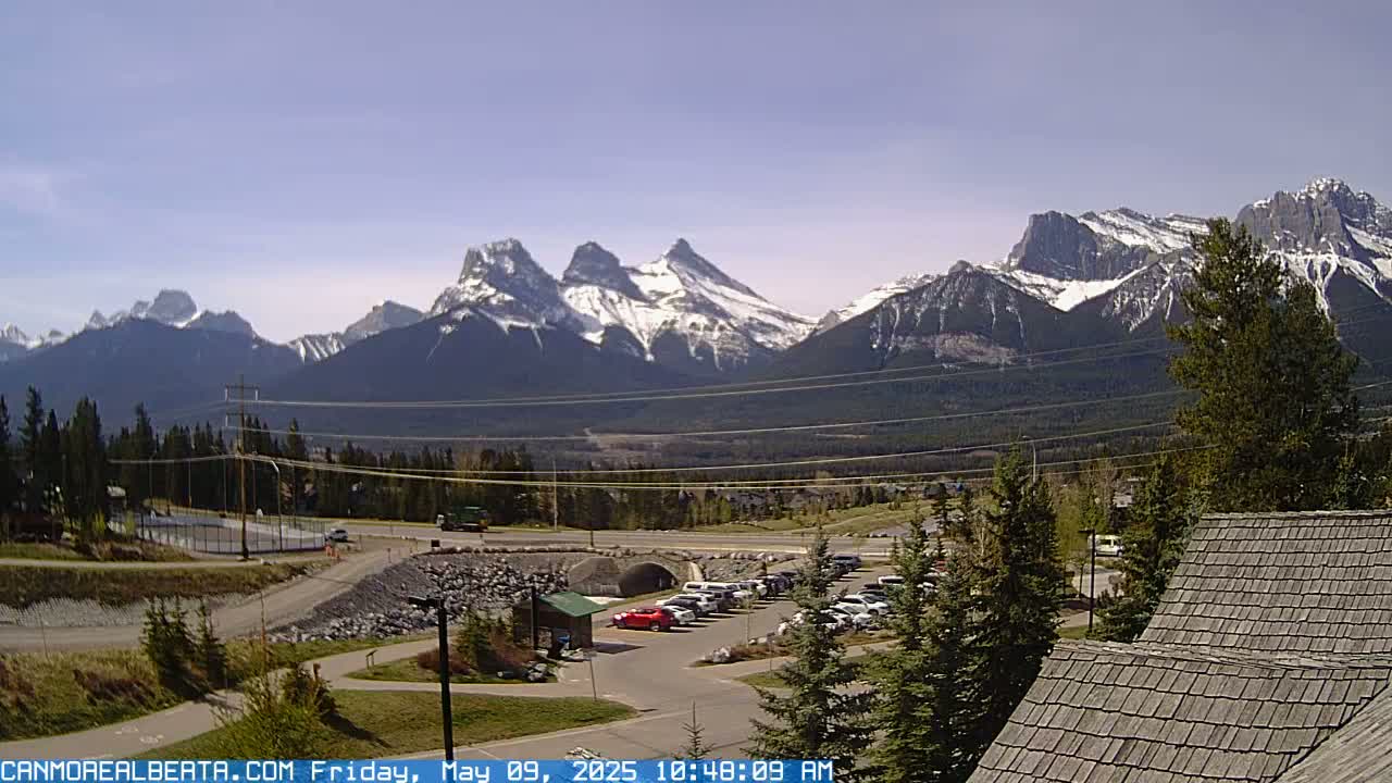 A parking lot and roadway are situated in the foreground, with snow-capped mountains stretching across the background under a clear sky.