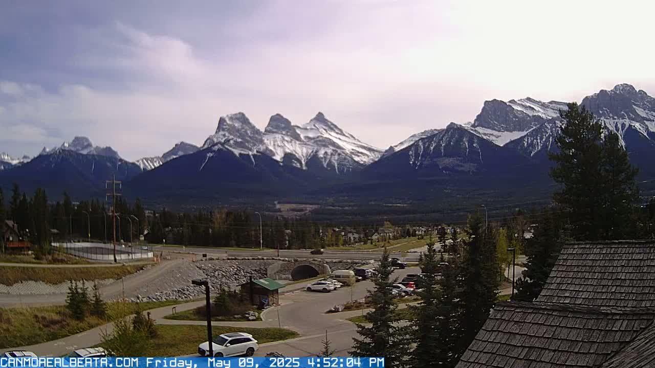 A mountain range capped with snow is visible in the distance under a partly cloudy sky, overlooking a parking lot and a small town.