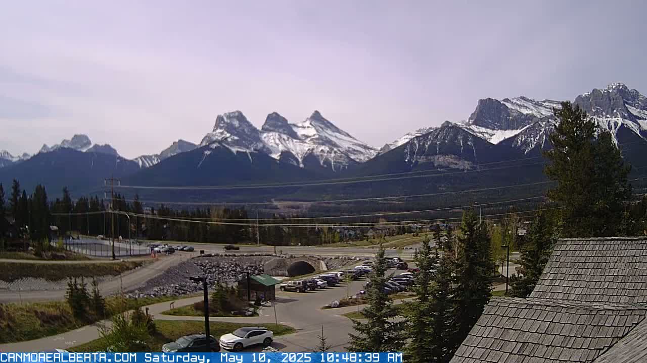 A parking lot with cars is situated in front of a mountain range partially covered in snow under a mostly clear sky.