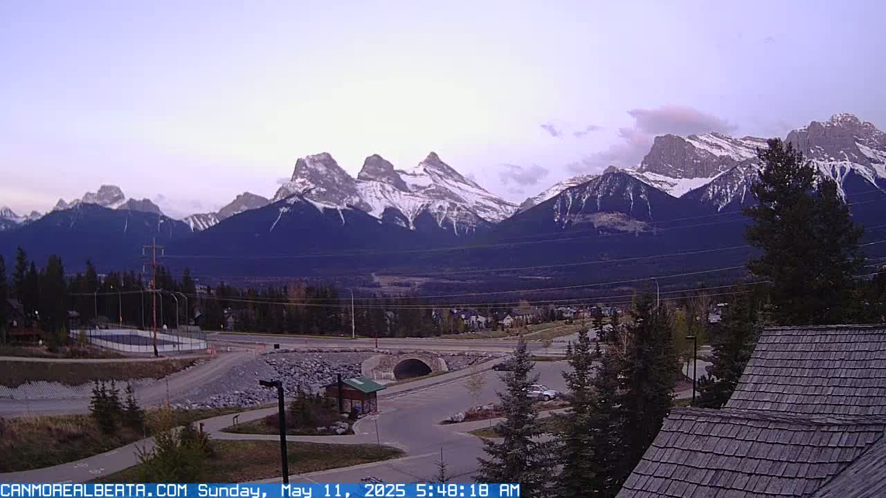 A view of snow-capped mountains under a partly cloudy dawn sky, overlooking a valley with roads, trees, and a small parking lot.