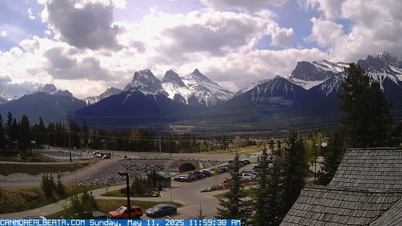 A partly cloudy day reveals snow-capped mountains in the distance, overlooking a valley with a parking lot, road, and a building with a shingled roof.
