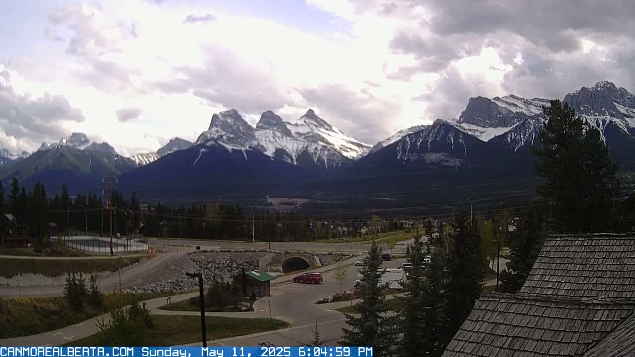 A partly cloudy day reveals snow-capped mountains in the distance, overlooking a valley with a parking lot, roadway, and some buildings.