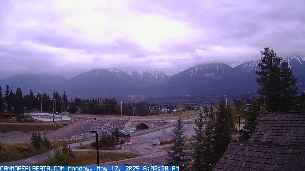 A partly cloudy, overcast day reveals snow-capped mountains in the distance, overlooking a valley with a road, a small bridge, and some buildings.