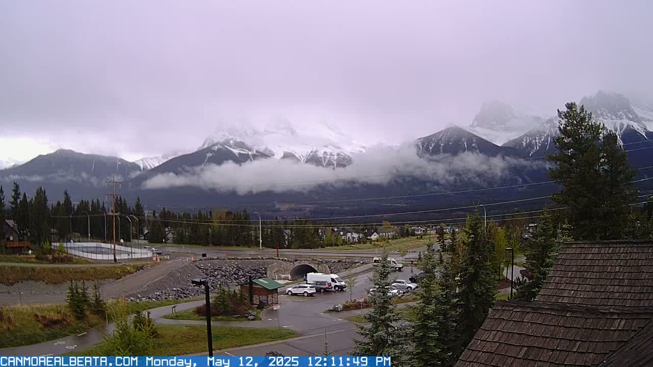 Snow-capped mountains are partially obscured by clouds over a valley town with several cars parked near a small building, under an overcast sky.