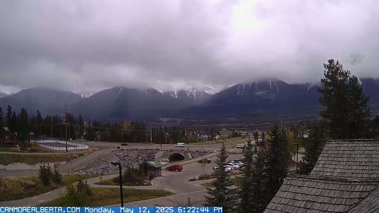 A cloudy day reveals snow-capped mountains in the distance, overlooking a valley with a road, buildings, and a parking lot.