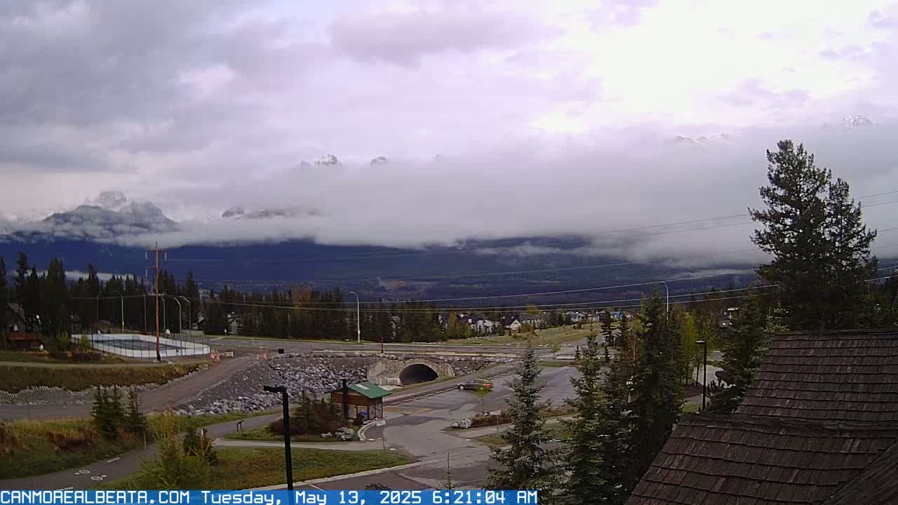 A cloudy day reveals mountains partially obscured by low-hanging clouds over a town with a parking lot and a small tunnel visible in the foreground.
