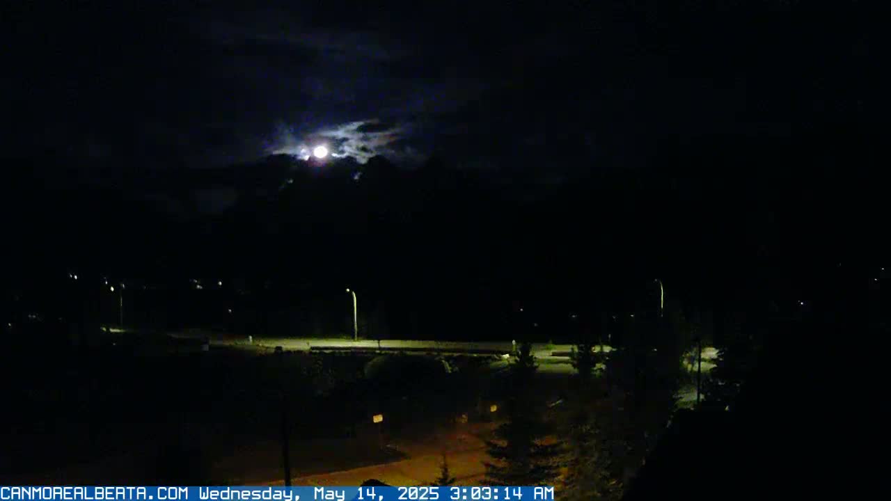 A bright moon is visible through partly cloudy night skies above a dark landscape with dimly lit roads and sparse vegetation.