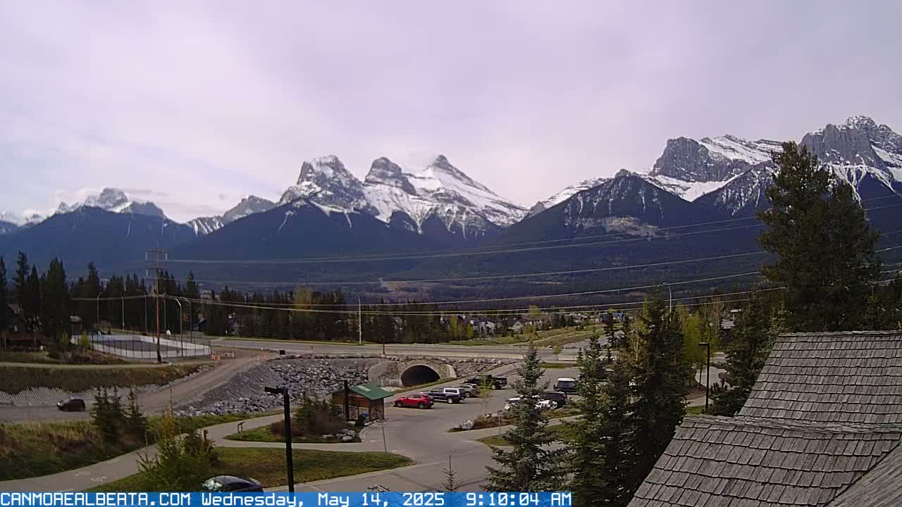 A partly cloudy day reveals snow-capped mountains in the distance, overlooking a valley with a roadway, parking lot, and some buildings.