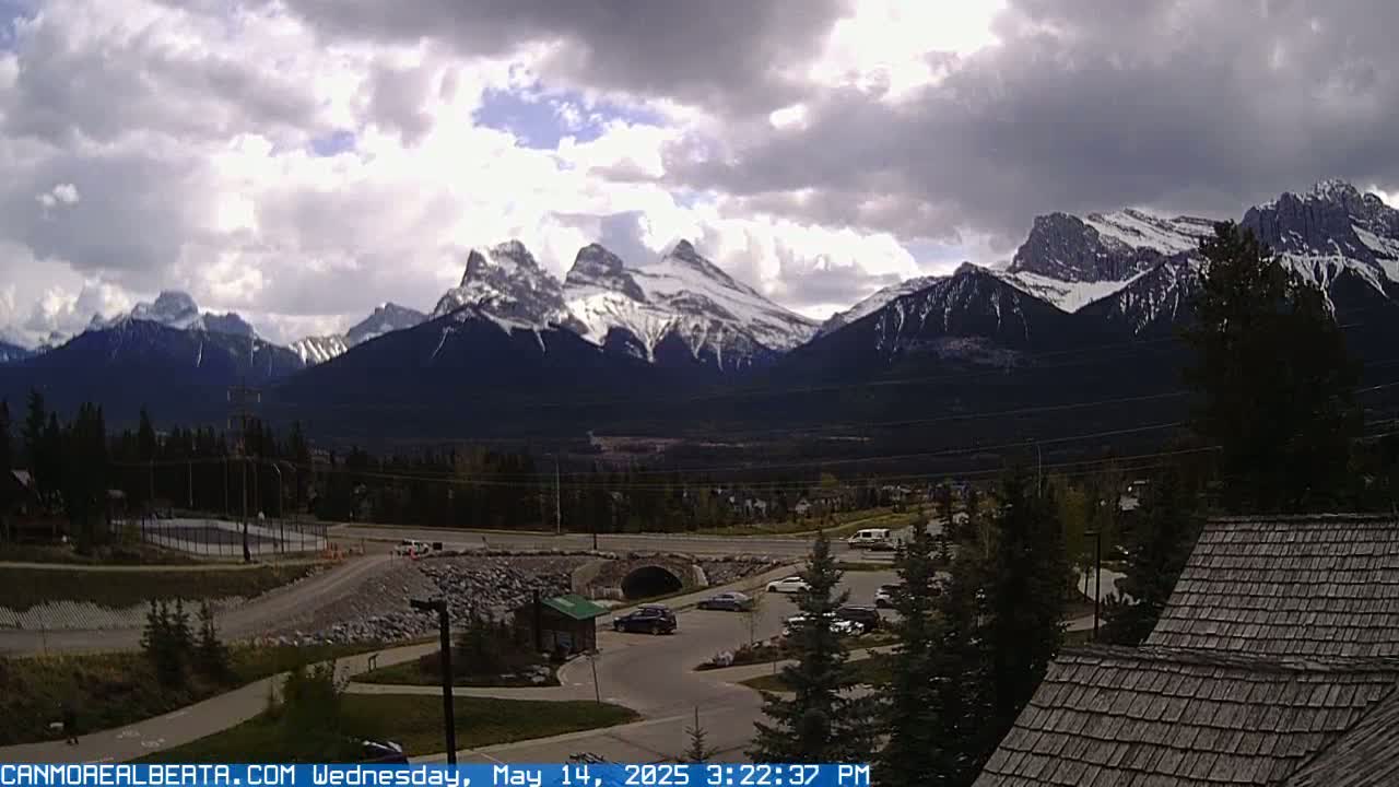 A partly cloudy day reveals snow-capped mountains in the distance, overlooking a parking lot, roadway, and a small building with a wood-shake roof.