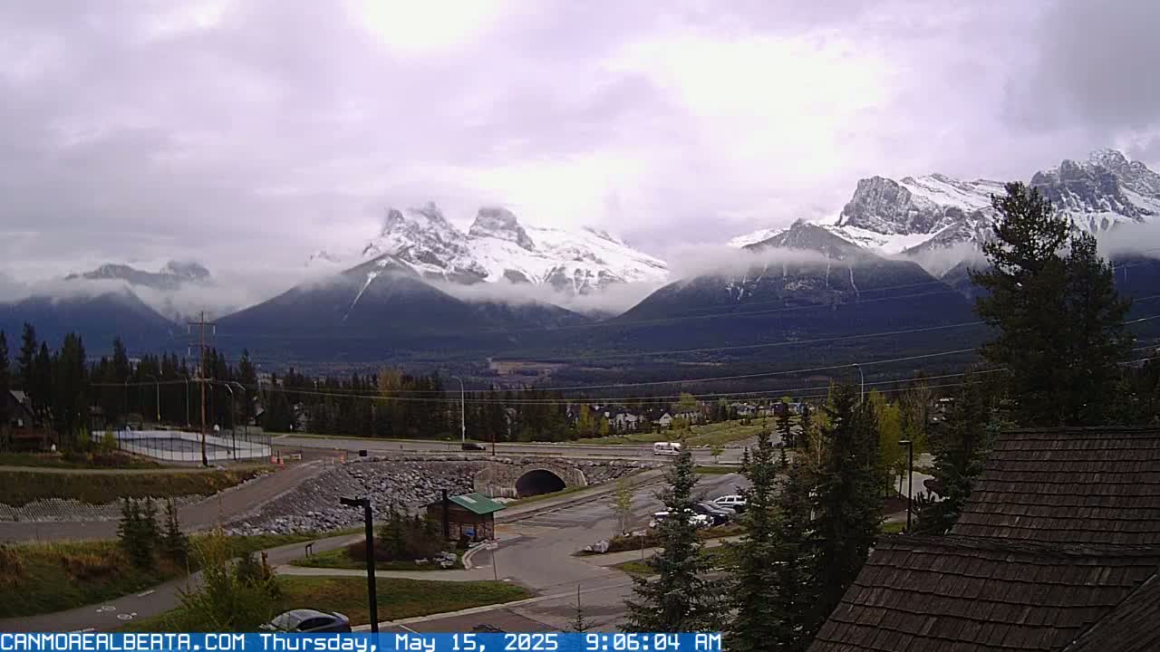A partly cloudy day reveals snow-capped mountains in the distance, overlooking a valley with a town, roads, and a parking lot.
