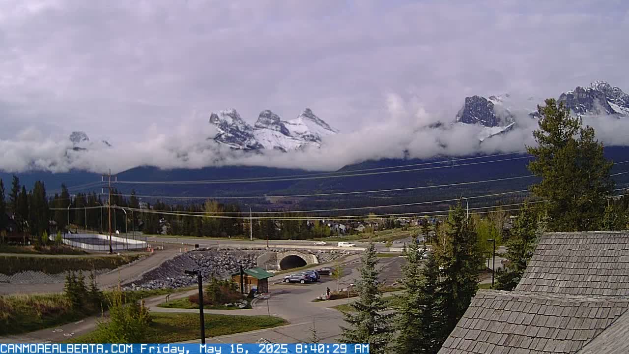 A partially cloudy day reveals snow-capped mountains in the distance, partially obscured by low-lying clouds, overlooking a valley with a roadway, parking lot, and some buildings.