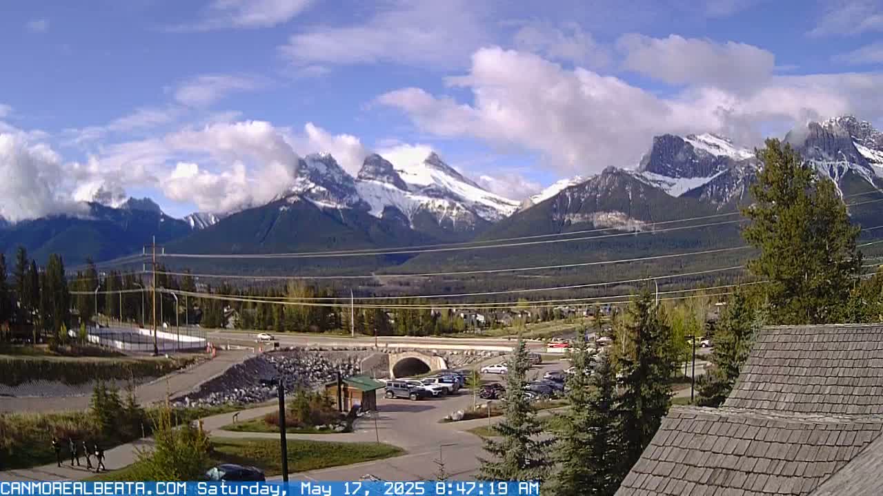 A partly cloudy day reveals snow-capped mountains in the distance, overlooking a valley with a road, bridge, parking lot, and some buildings.