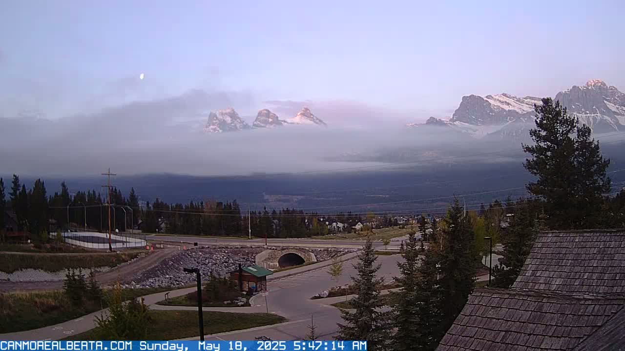 Snow-capped mountains partially obscured by fog are visible in the distance, overlooking a paved road, parking lot, and some buildings under a partly cloudy, dusky sky.