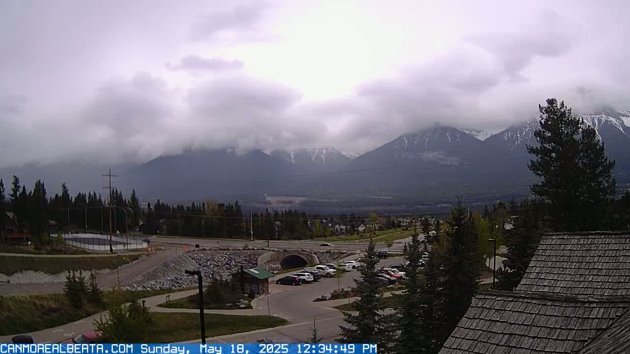 A cloudy day reveals a mountain range in the distance, with a parking lot, road, and some buildings in the foreground.