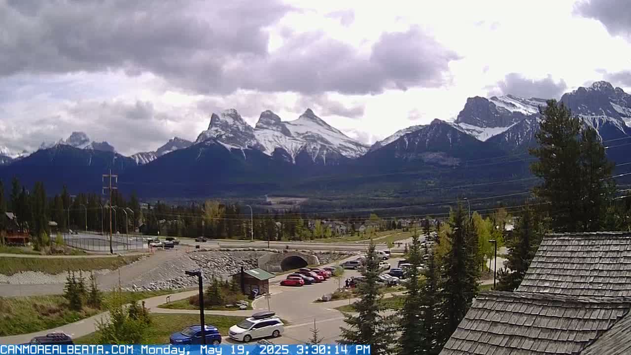 A parking lot with several cars is situated in front of a valley, with snow-capped mountains in the background under a partly cloudy sky.