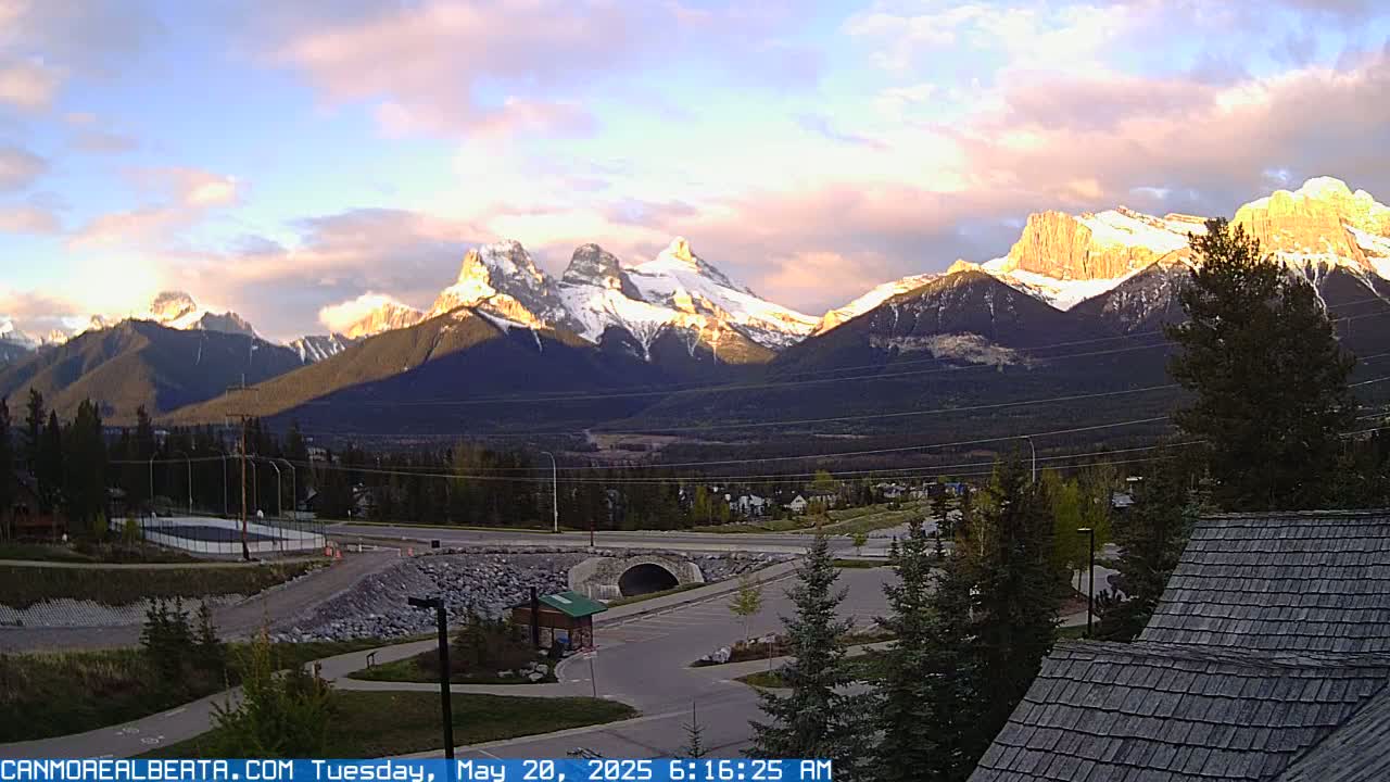 A sunrise illuminates snow-capped mountains in the distance, overlooking a parking lot, a small bridge, and some buildings.