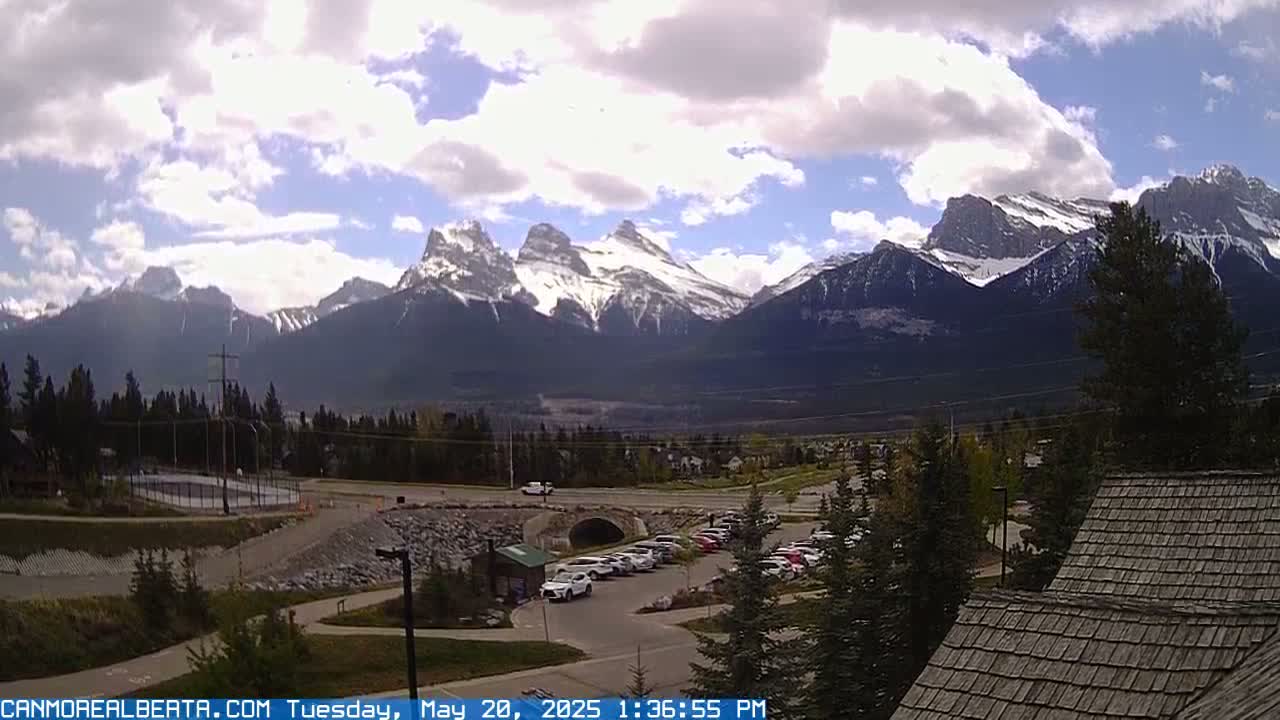 A partly cloudy day reveals snow-capped mountains in the distance, overlooking a parking lot and a residential area with evergreen trees.