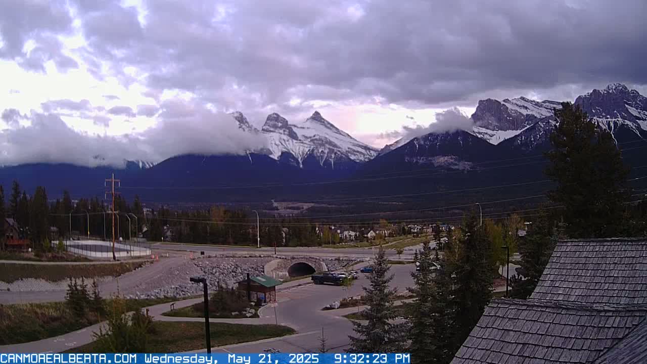 A partly cloudy sky hangs over snow-capped mountains, a town, and a parking lot; a portion of a building's roof is visible in the bottom right corner.