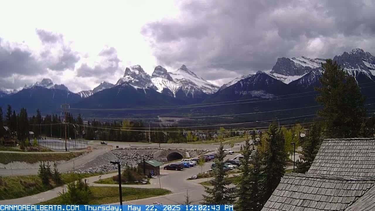 A parking lot with cars sits in front of a partially cloudy sky and a mountain range with snow-capped peaks in the background.