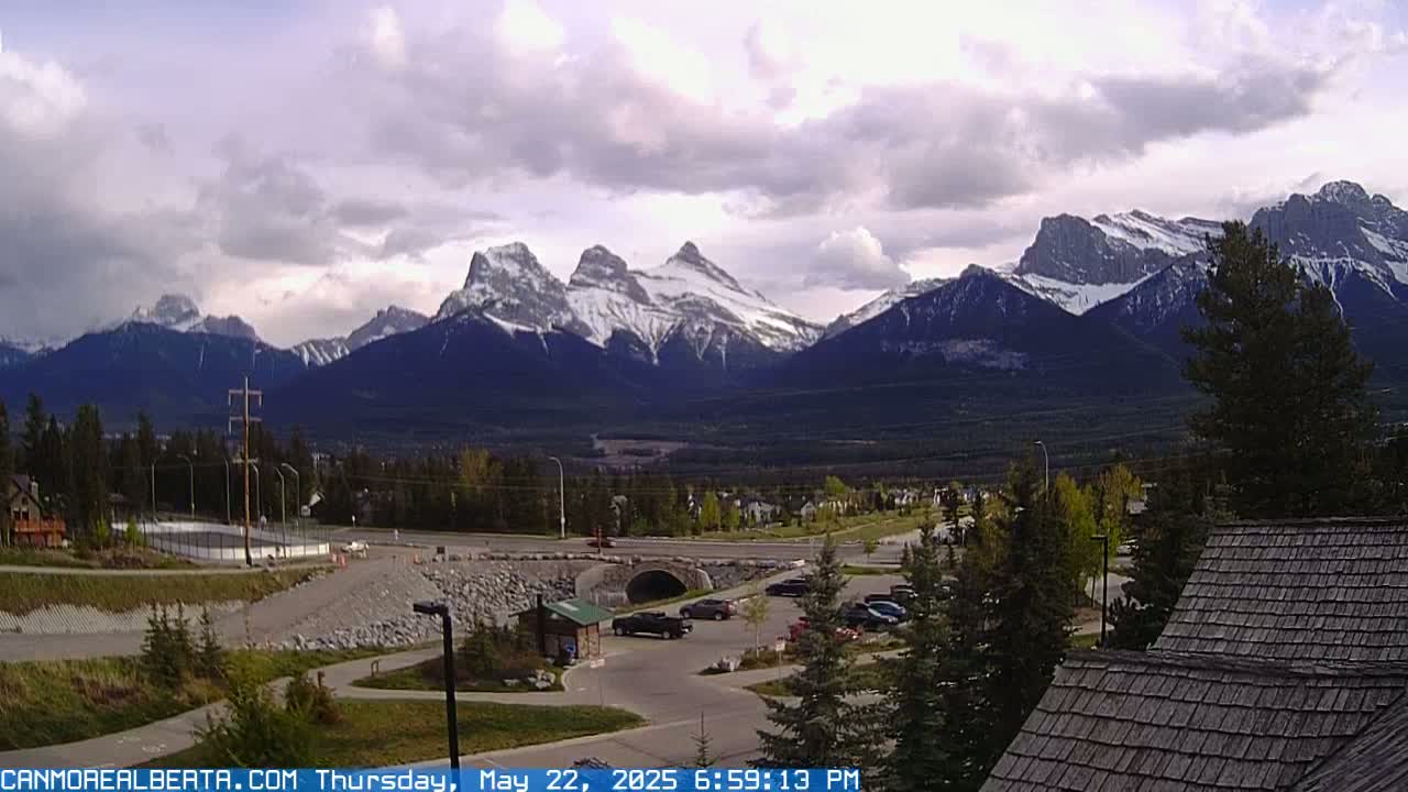 A partially cloudy sky overlooks a valley with a roadway, parking lot, and some buildings, all set against a backdrop of snow-capped mountains.