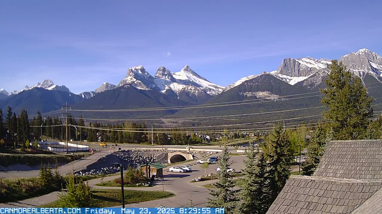 A clear, sunny day reveals snow-capped mountains in the distance, overlooking a small town with a parking lot, a road, and a tunnel, partially obscured by power lines and trees.
